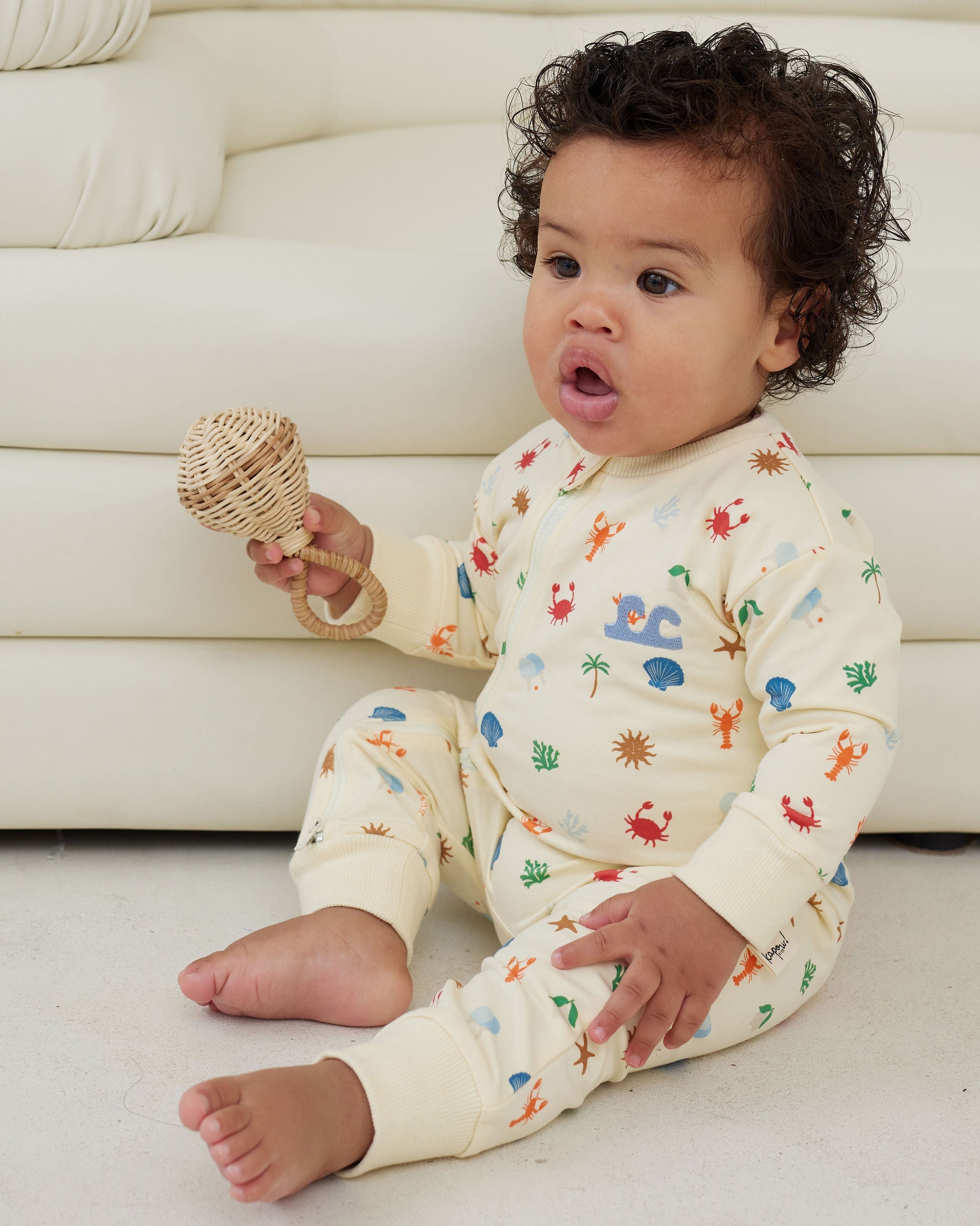 Baby wearing a cream-colored onesie with colorful patterns, sitting on a white floor against a white couch.