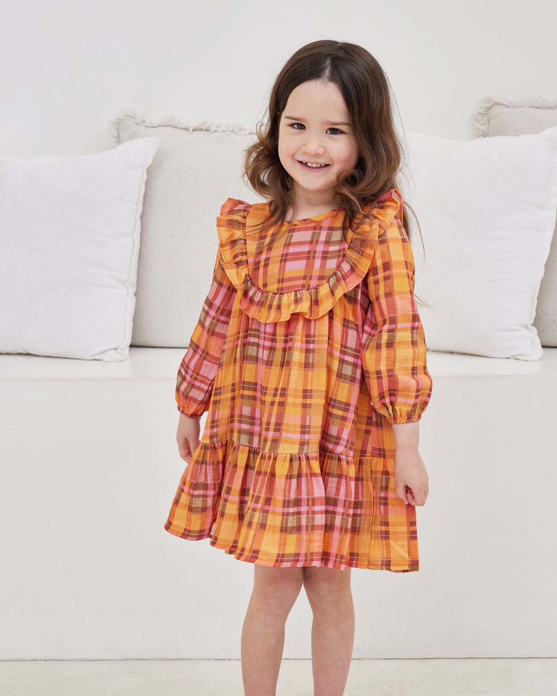 Young girl wearing a plaid dress standing in front of a white couch.